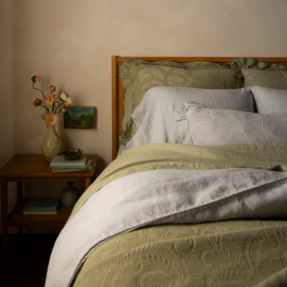 Eye-level shot of a bed against a wood and cane headboard, dressed with desert sage Marisette shams, cloud Linen pillowcases, a cloud Marisette throw pillow, desert sage Linen sheets, a cloud Linen duvet cover, and a desert sage Marisette bedspread draped over the bed