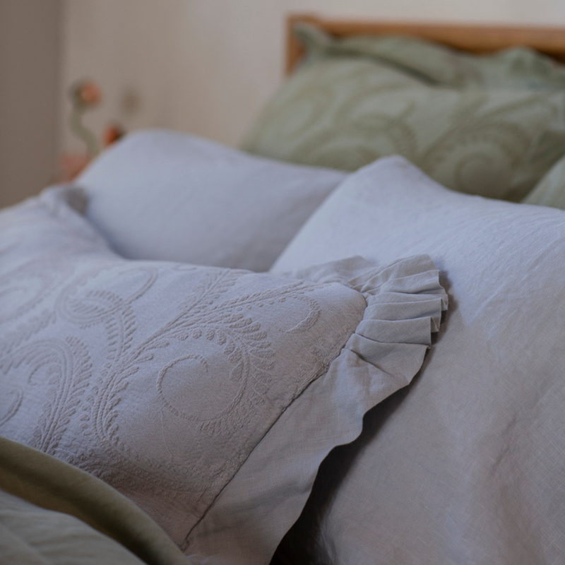 Marisette Throw Pillow | a close up of an organic cotton damask 16x36-inch throw pillow in cloud showing the gathered ruffle at the corner against a sleeping pillow with shams out of focus in the background.