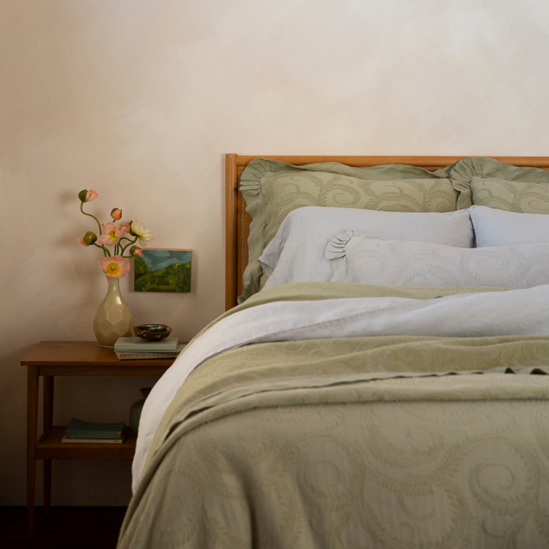 Marisette Bedspread | a bed dressed in light blue and green tones with a rattan headboard and matching side table - shown from the foot of the bed.