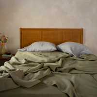 A unmade bed against a wood and cane headboard, dressed with desert sage Linen sheets and cloud Linen pillowcases, shown in a straight-on, eye-level shot