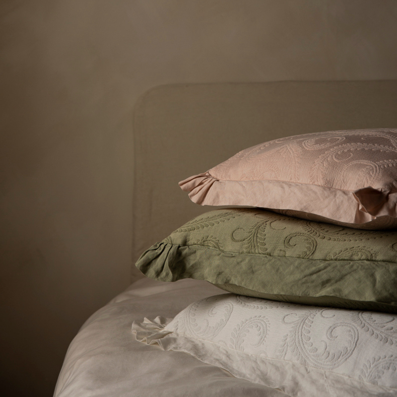 Marisette Throw Pillow | a stack of organic cotton damask throw pillows on a bed with moody lighting;  pillows are pink, green and white tones.