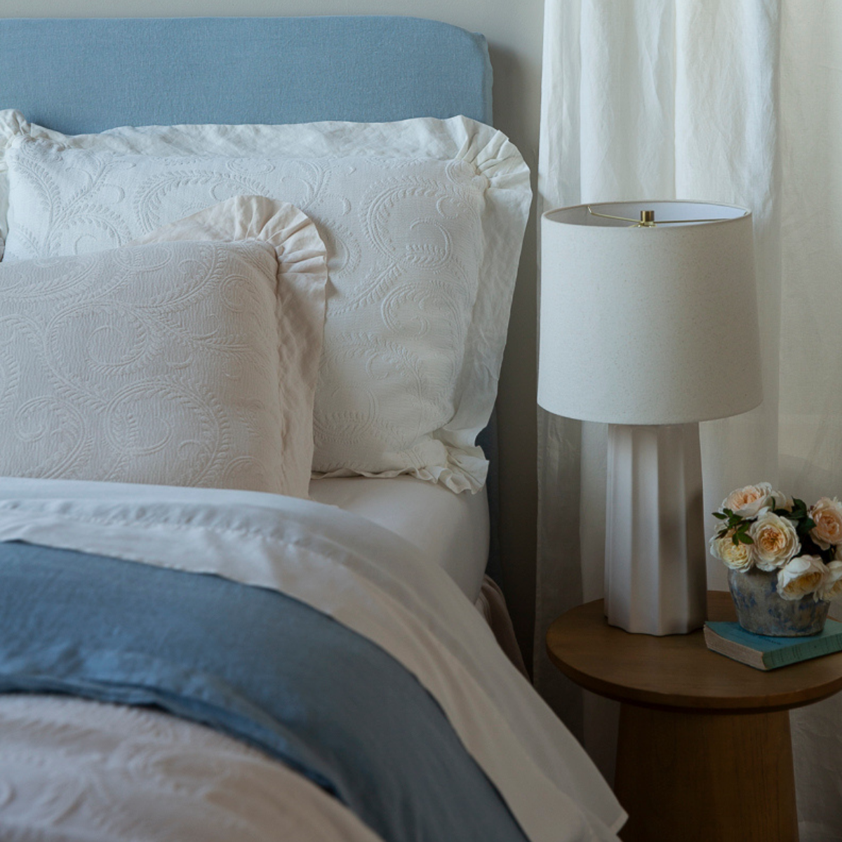 Marisette Sham | organic cotton damask trimmed in linen shams against a blue headboard with blue and white bedding, a lamp and small bouquet of flowers on the side teble.