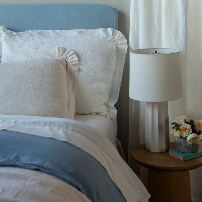 Marisette Sham | organic cotton damask trimmed in linen shams against a blue headboard with blue and white bedding, a lamp and small bouquet of flowers on the side teble.