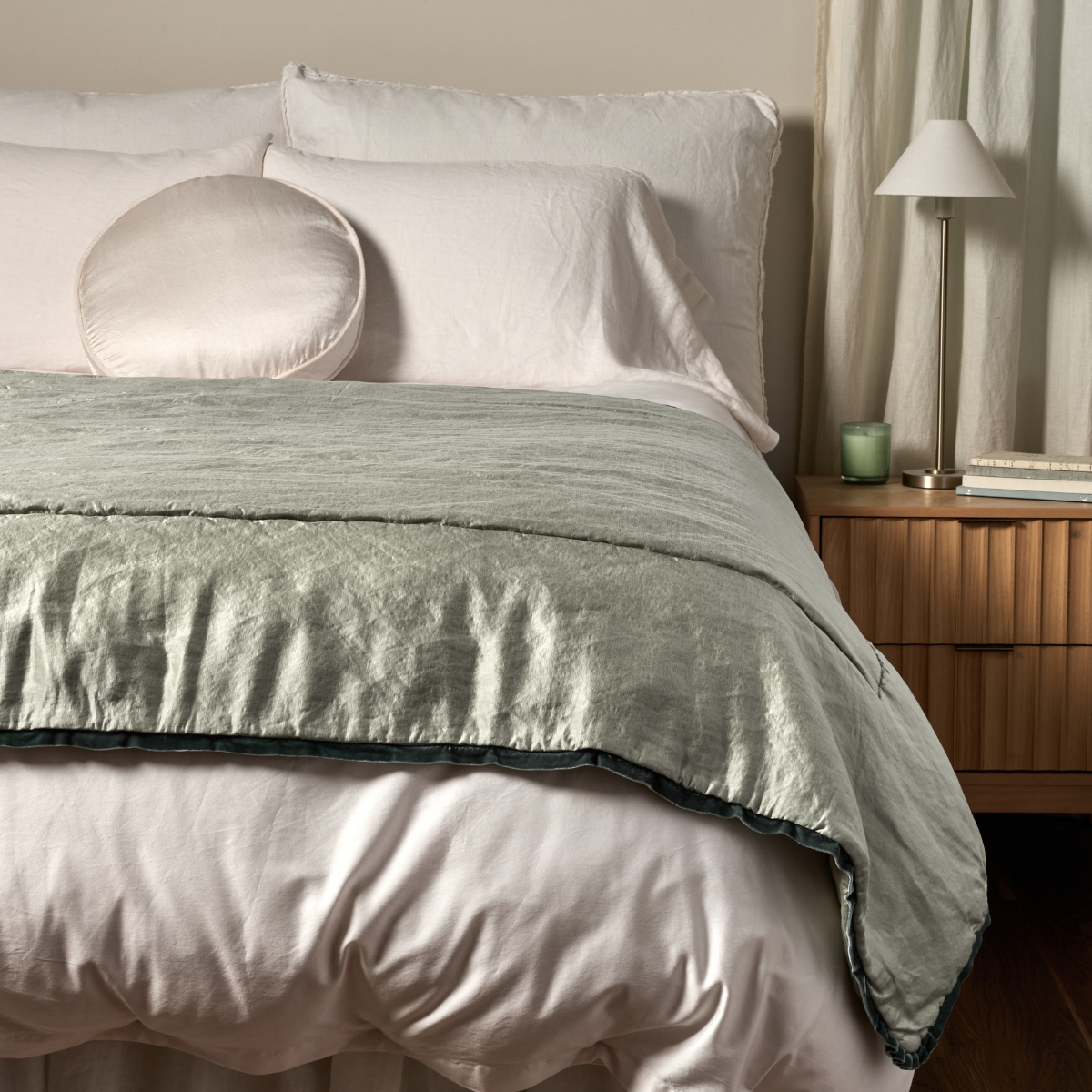 Paloma Blanket | Eucalyptus | a charmeuse bed end blanket is shown layered at the foot of a bed, its contrasting silk velvet trim and petit ruffle at the corner visible against the all winter white bedding.