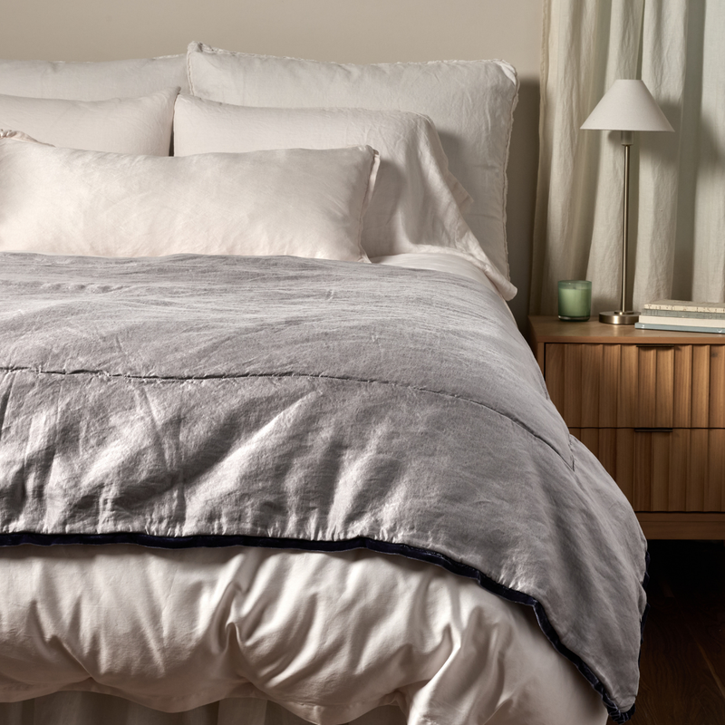 Paloma Blanket | French Lavender | a charmeuse bed end blanket is shown layered at the foot of a bed, its contrasting trim and petit ruffle at the corner visible against the all winter white bedding.