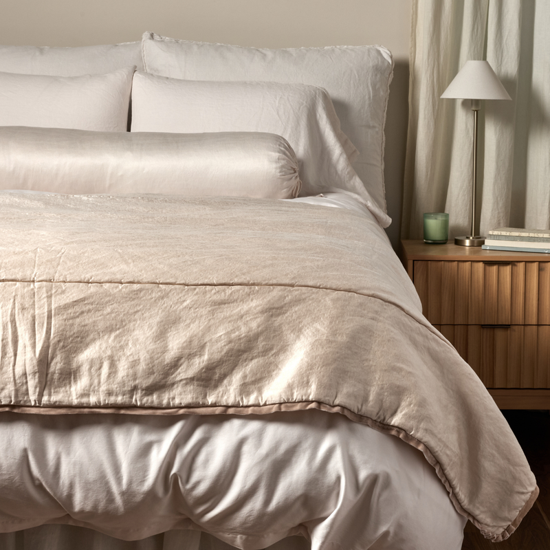 Paloma Blanket | Pearl | a charmeuse bed end blanket is shown layered at the foot of a bed, its contrasting silk velvet trim and petit ruffle at the corner visible against the all winter white bedding.