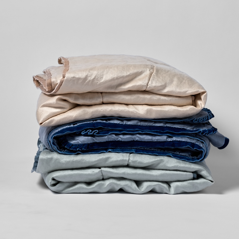 Paloma Blanket | a stack of three folded charmeuse blankets in pearl, tahoe blue, and cloud shown against a white background.