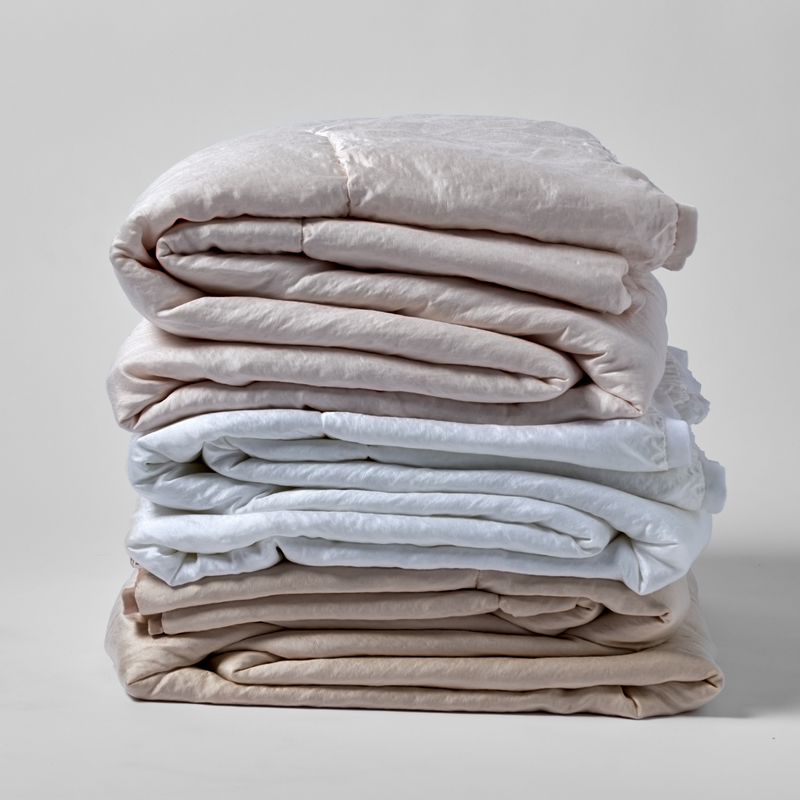 Paloma Blanket | a stack of three folded charmeuse blankets in blush, white, and pearl shown against a white background. 