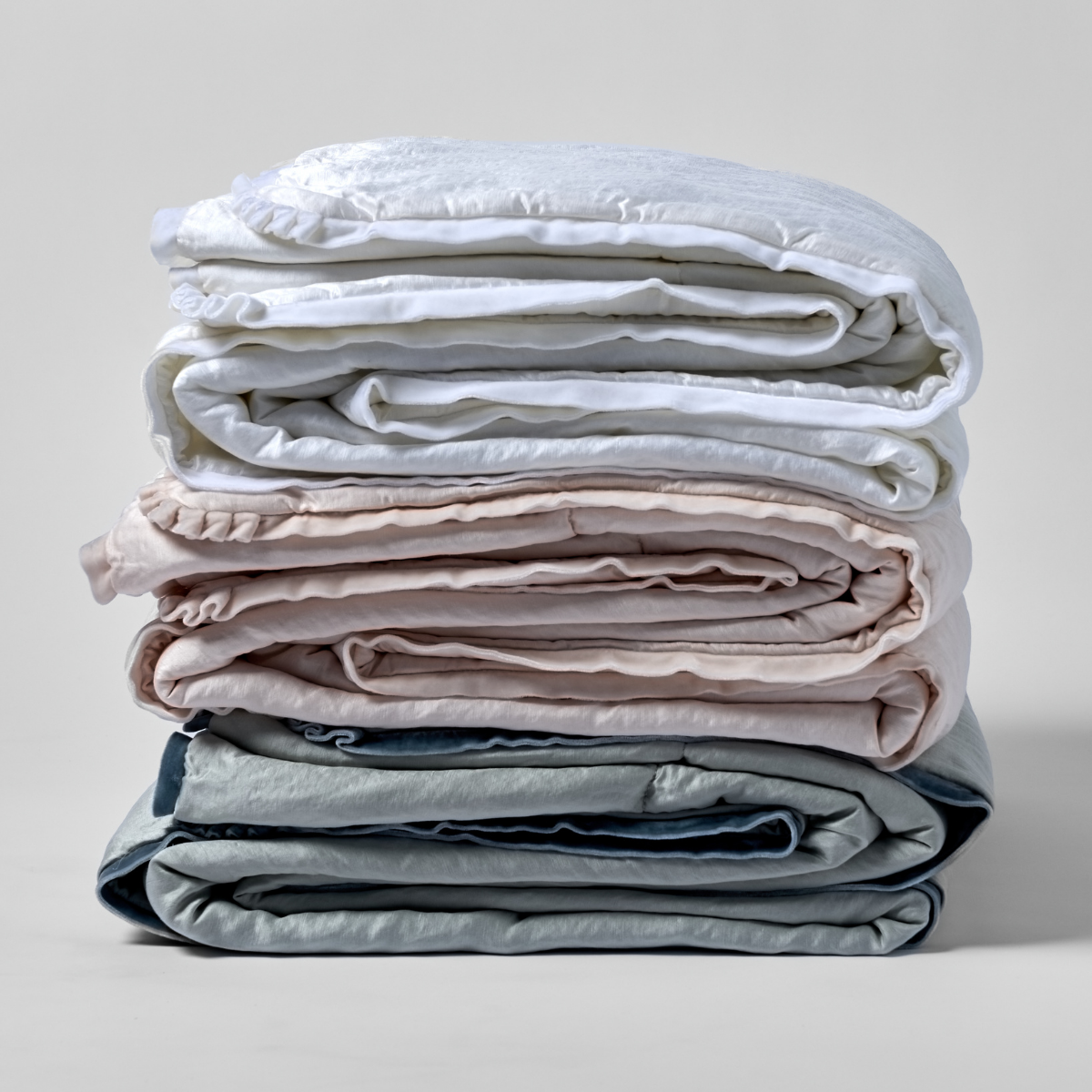 Paloma Blanket | a stack of three folded charmeuse blankets in white, pearl, and cloud shown against a white background. 