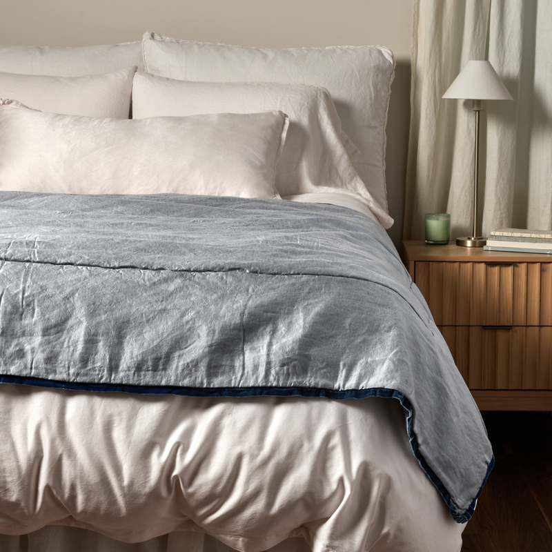 Paloma Blanket | Tahoe Blue | a charmeuse bed end blanket is shown layered at the foot of a bed, its contrasting silk velvet trim and petit ruffle at the corner visible against the all winter white bedding.