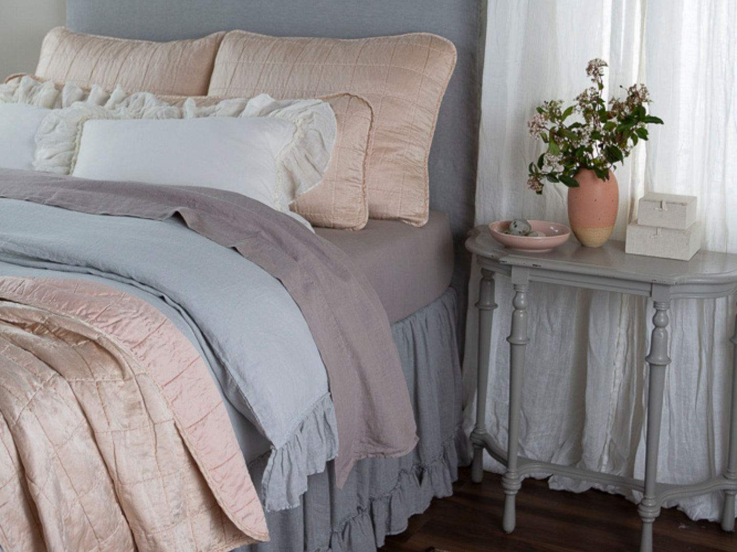 A corner shot of a bed dressed in a soft palette and delicate ruffles unite with the structure of our square-quilt collection for a modern farmhouse look to best suit your serenity sanctuary. A gray table with decor of a similar palette sits next to the bed.