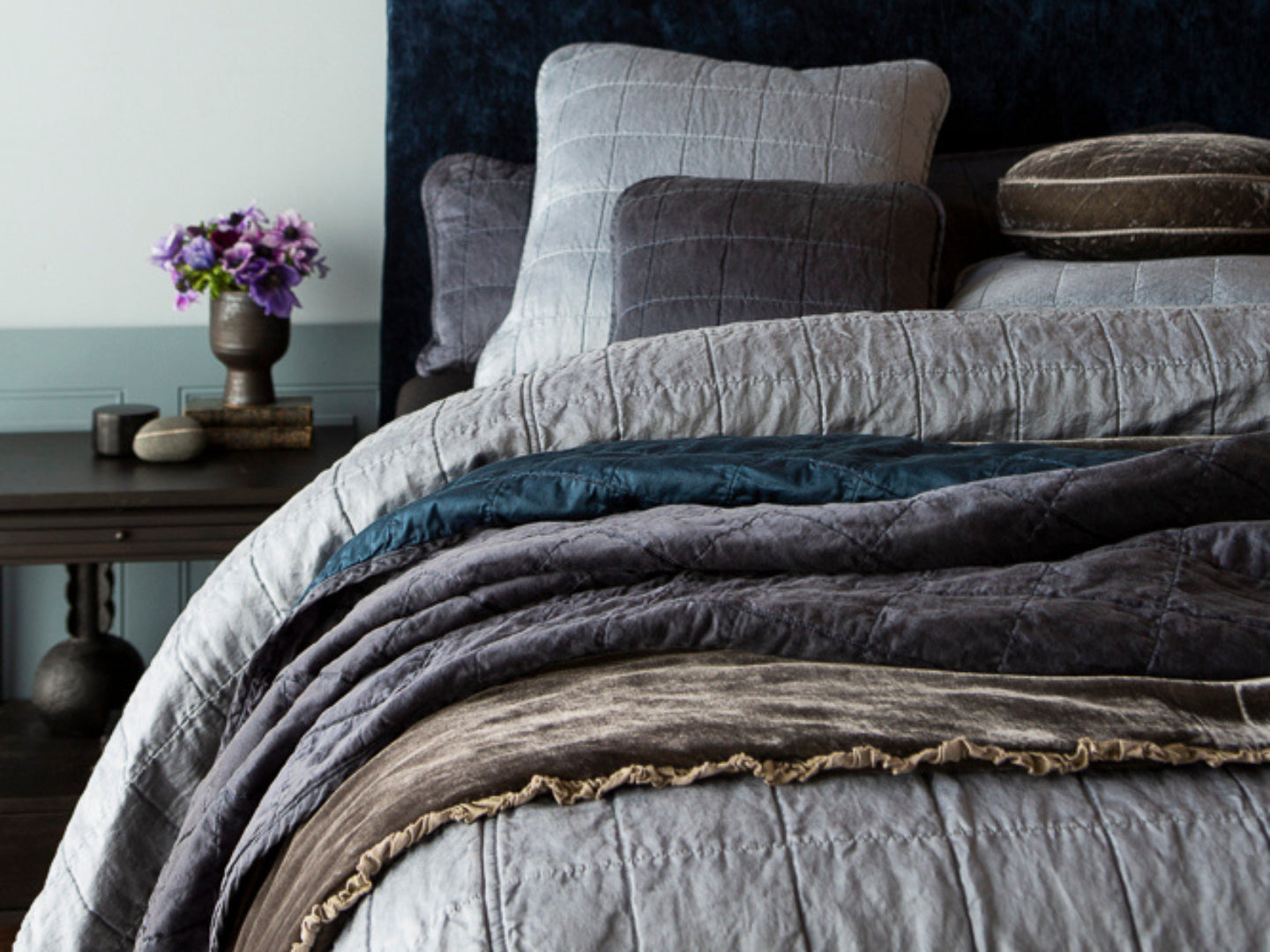 The new Sel collection is front and center in a moody palette providing a contemporary look. A dark wooden table adorned with a vibrant purple flower arrangement sits next to the bed. 