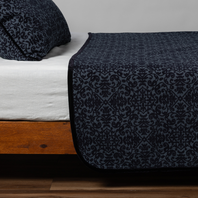 Slate | Matching Vienna Coverlet and Sham  in slate, a polished dusky dark blue, shown as the sideview on a bed against a plain background.