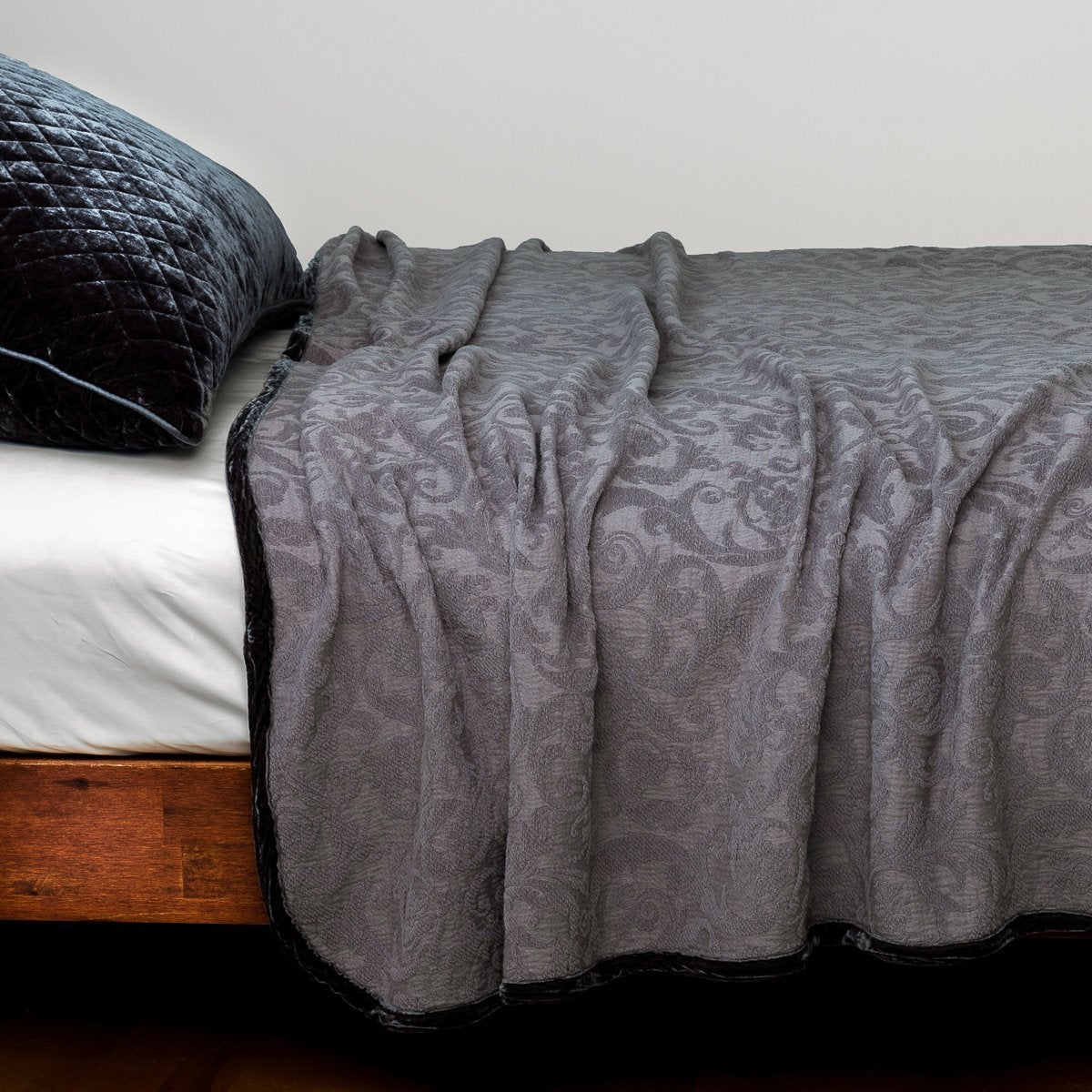 Adele Twin Coverlet | The Adele coverlet in moonlight, styled on a wood framed bed shown from the side. Shown with a white sheet and a silk velvet quilted sham in moonlight.