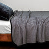 The Adele coverlet in moonlight, styled on a wood framed bed shown from the side. Shown with a white sheet and a silk velvet quilted sham in moonlight.