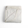 Adele Blanket | Winter White | folded organic cotton damask throw blanket shown with the corner folded down to show silk velvet trim detail — overhead against a plain white background.