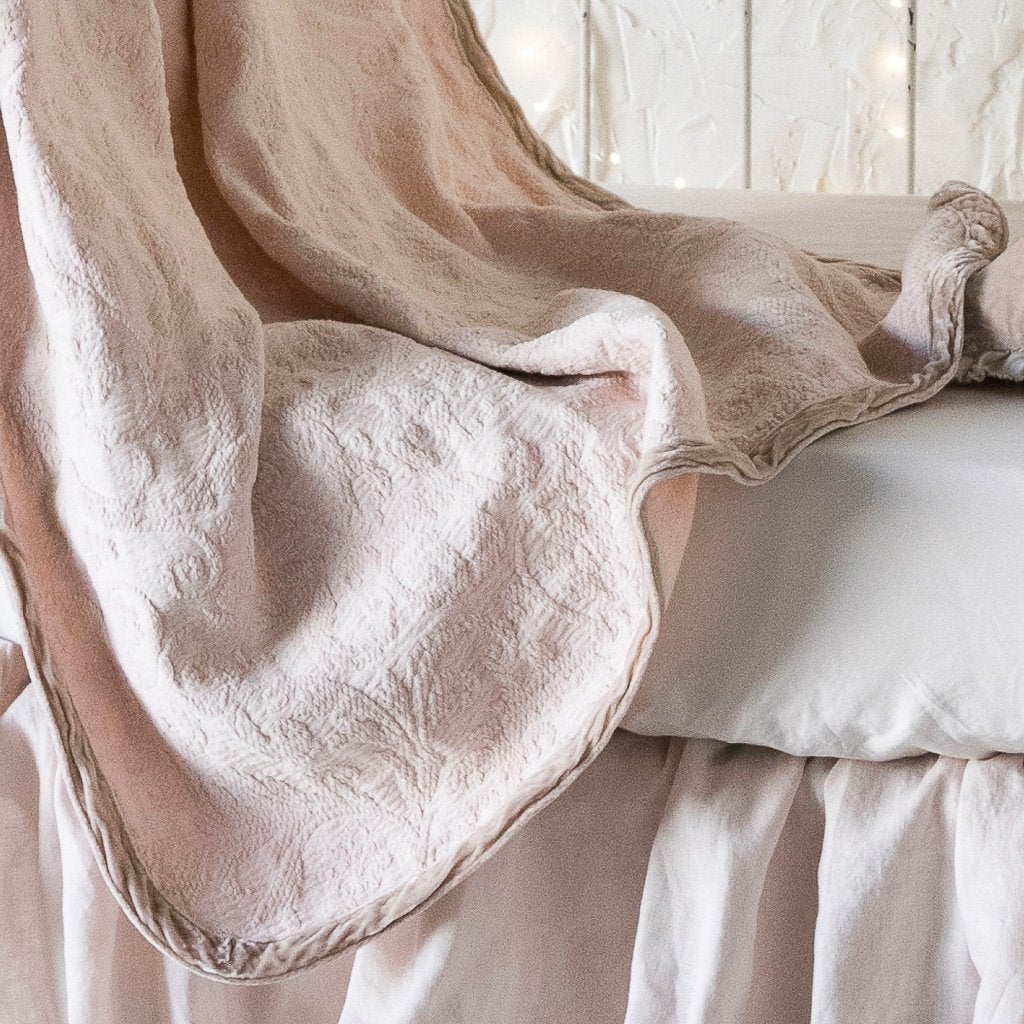 Pearl | A close up of the Adele baby blanket in pearl draped over a crib mattress, highlighting the texture of the cotton damask and the silk velvet edging, which absorbs the dye more richly.