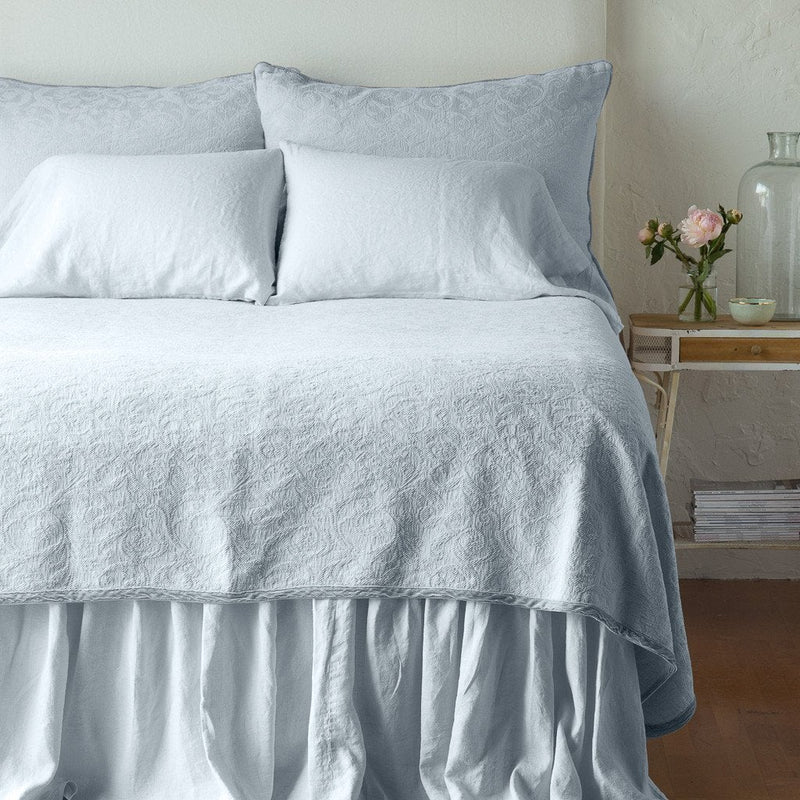 Cloud | The Adele coverlet in cloud, viewed from the foot of the bed, with white bed skirt and pillows visible. The headboard is plain light color, and part of a bedside table is visible with flowers, a vase and some books. against a plain white wall.