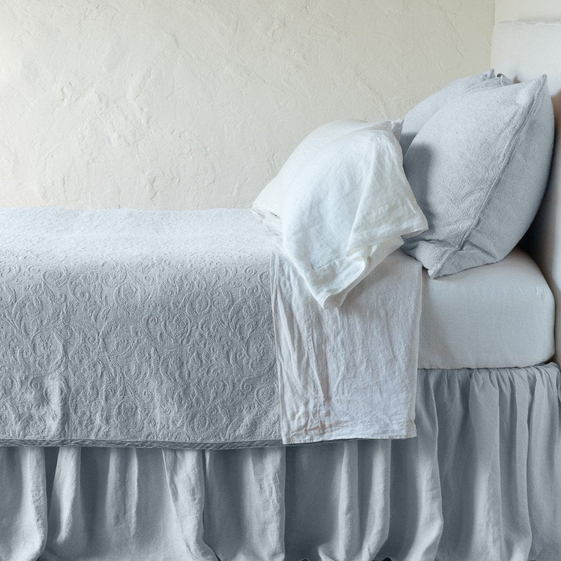 Cloud | The Adele coverlet in cloud, on a bed viewed from the side against a plain white wall. The bed is styled neatly with the coverlet folded back to reveal white sheets, bed skirt and pillows.