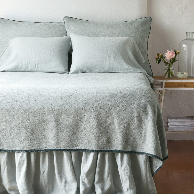 Eucalyptus | The Adele coverlet in eucalyptus, viewed from the foot of the bed, with white bed skirt and pillows visible. The headboard is plain light color, and part of a bedside table is visible with flowers, a vase and some books. against a plain white wall.