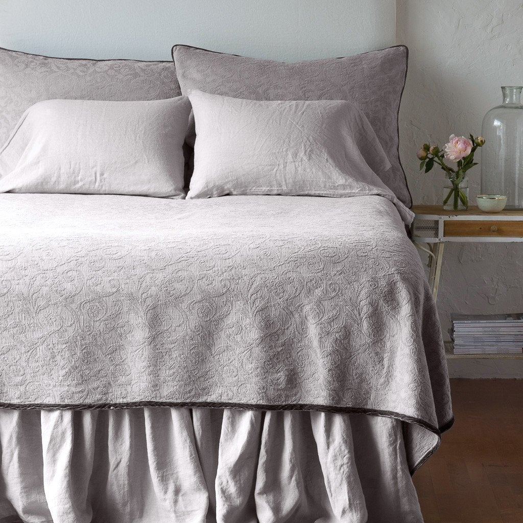 Fog | The Adele coverlet in fog, viewed from the foot of the bed, with white bed skirt and pillows visible. The headboard is plain light color, and part of a bedside table is visible with flowers, a vase and some books. against a plain white wall.