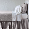 Fog | The Adele coverlet in fog, on a bed viewed from the side against a plain white wall. The bed is styled neatly with the coverlet folded back to reveal white sheets, bed skirt and pillows.