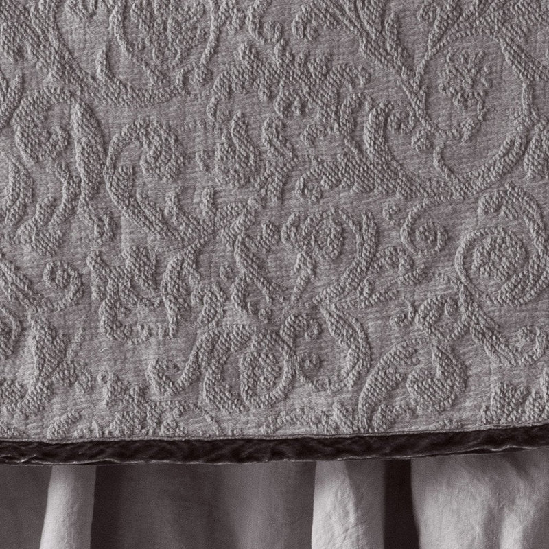 Fog | Close up of the Adele coverlet in fog, highlighting the detail of the cotton damask and the silk velvet edging trim. A white bed skirt peeks out at the bottom of the shot.