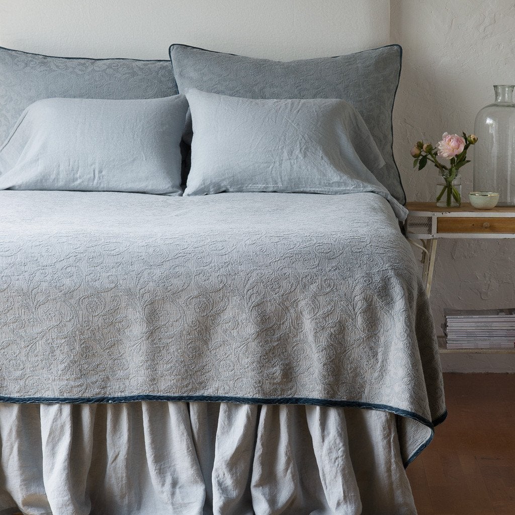 Mineral | The Adele coverlet in mineral, viewed from the foot of the bed, with white bed skirt and pillows visible. The headboard is plain light color, and part of a bedside table is visible with flowers, a vase and some books. against a plain white wall.