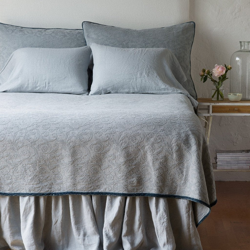 Mineral | The Adele coverlet in mineral, viewed from the foot of the bed, with white bed skirt and pillows visible. The headboard is plain light color, and part of a bedside table is visible with flowers, a vase and some books. against a plain white wall.