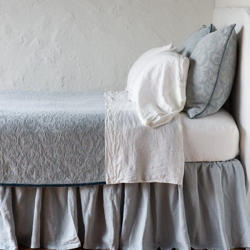Mineral | The Adele coverlet in mineral, on a bed viewed from the side against a plain white wall. The bed is styled neatly with the coverlet folded back to reveal white sheets, bed skirt and pillows.