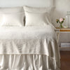 Parchment | The Adele coverlet in parchment, viewed from the foot of the bed, with parchment bed skirt and pillows visible. The headboard is plain light color, and part of a bedside table is visible with flowers, a vase and some books. against a plain white wall.