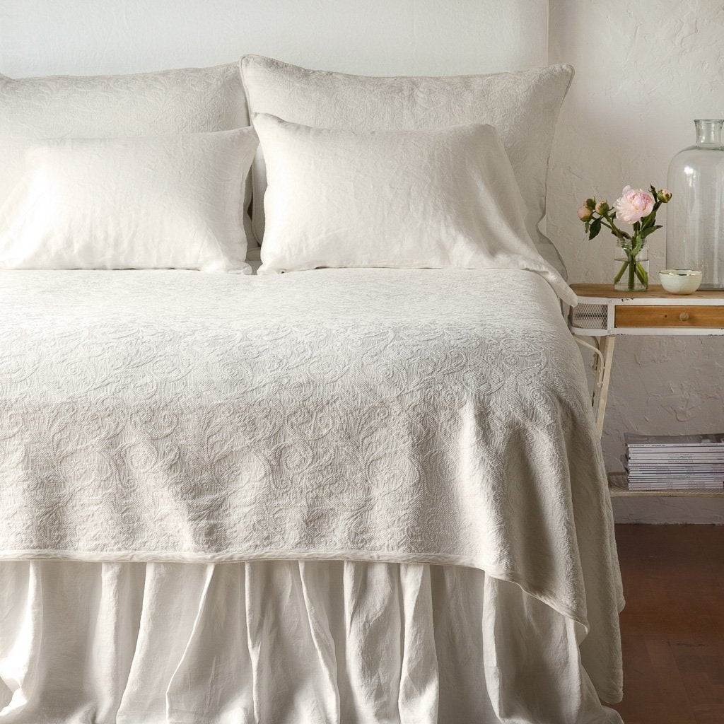 Parchment | The Adele coverlet in parchment, viewed from the foot of the bed, with parchment bed skirt and pillows visible. The headboard is plain light color, and part of a bedside table is visible with flowers, a vase and some books. against a plain white wall.