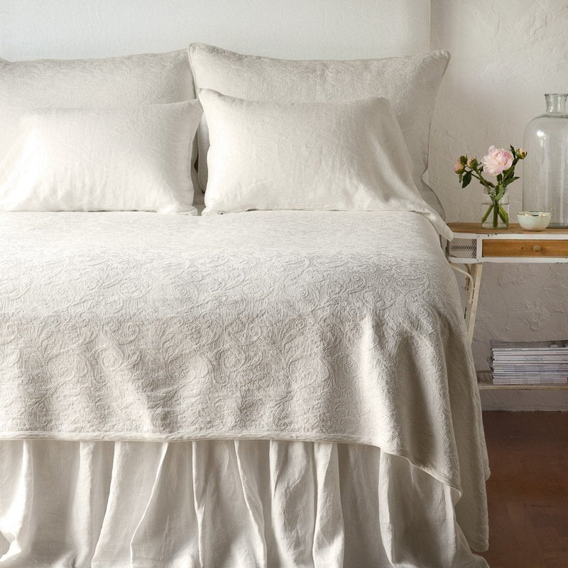 Parchment | The Adele coverlet in parchment, viewed from the foot of the bed, with parchment bed skirt and pillows visible. The headboard is plain light color, and part of a bedside table is visible with flowers, a vase and some books. against a plain white wall.