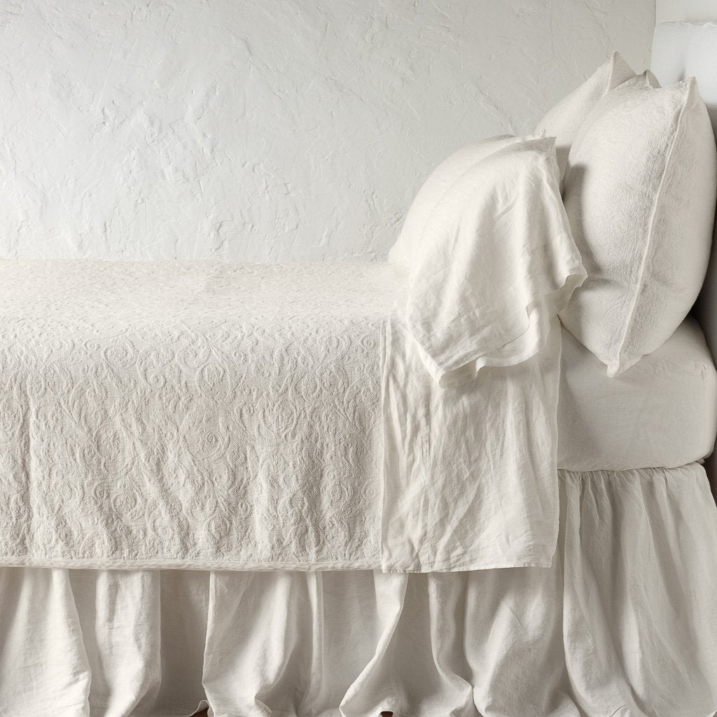Parchment | The Adele coverlet in parchment, on a bed viewed from the side against a plain white wall. The bed is styled neatly with the coverlet folded back to reveal parchment sheets, bed skirt and pillows.