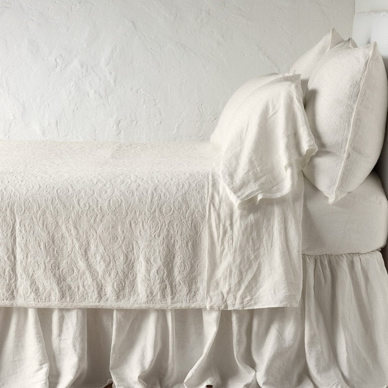 Parchment | The Adele coverlet in parchment, on a bed viewed from the side against a plain white wall. The bed is styled neatly with the coverlet folded back to reveal parchment sheets, bed skirt and pillows.