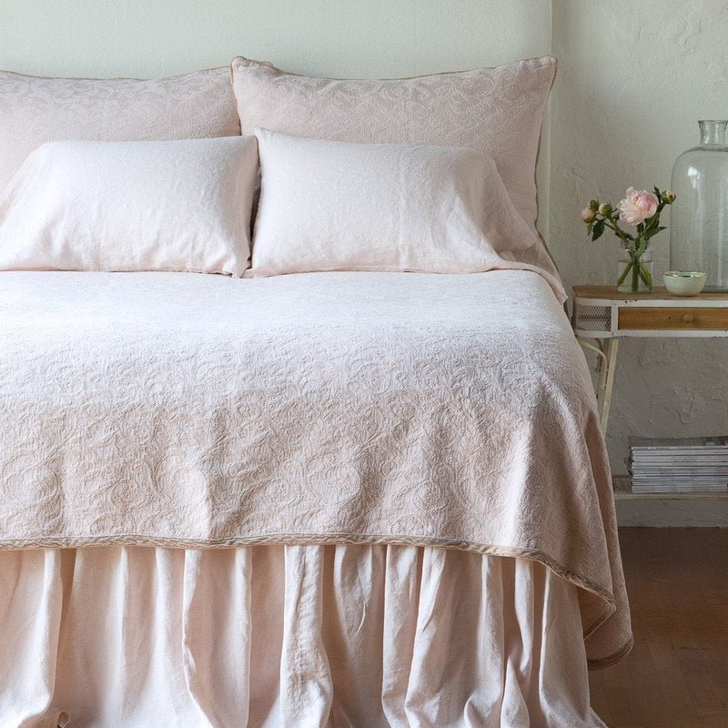 Pearl | The Adele coverlet in pearl, viewed from the foot of the bed, with white bed skirt and pillows visible. The headboard is plain light color, and part of a bedside table is visible with flowers, a vase and some books. against a plain white wall.