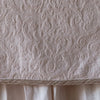 Pearl | Close up of the Adele coverlet in pearl, highlighting the detail of the cotton damask and the silk velvet edging trim. A white bed skirt peeks out at the bottom of the shot.