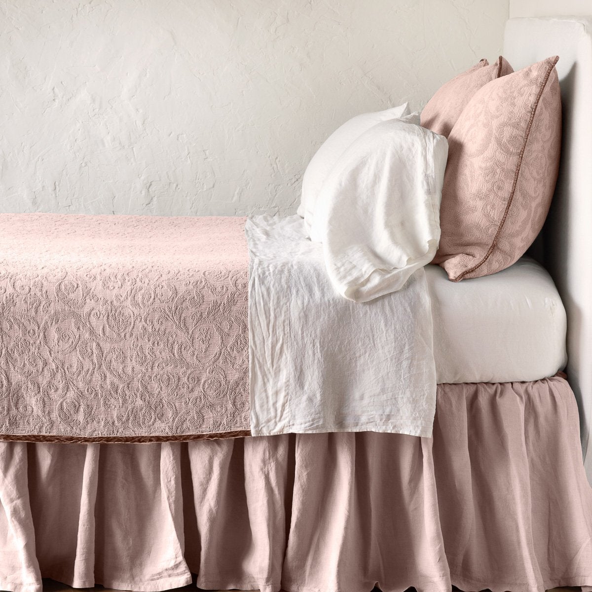 Rouge | The Adele coverlet in rouge, on a bed viewed from the side against a plain white wall. The bed is styled neatly with the coverlet folded back to reveal white sheets, bed skirt and pillows.