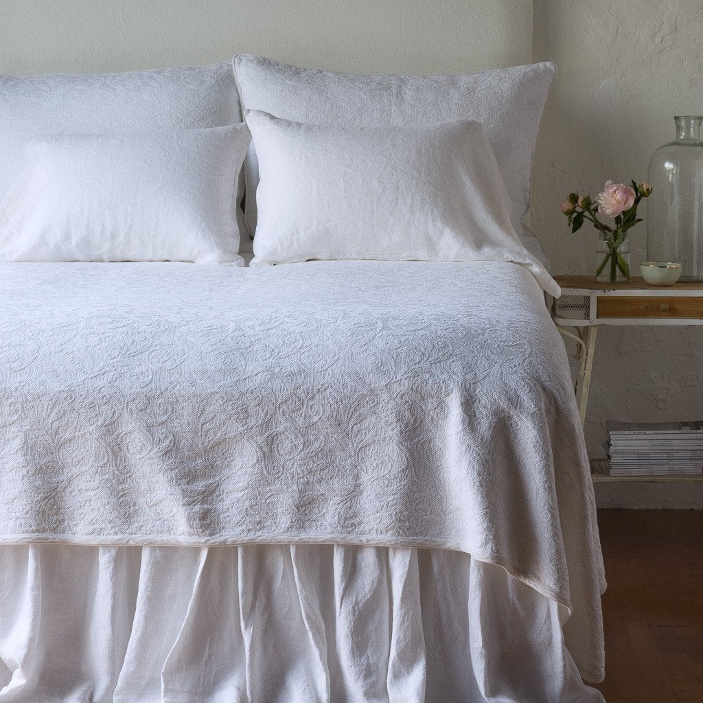 White | The Adele coverlet in white, viewed from the foot of the bed, with white bed skirt and pillows visible. The headboard is plain light color, and part of a bedside table is visible with flowers, a vase and some books. against a plain white wall.