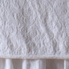 White | Close up of the Adele coverlet in white, highlighting the detail of the cotton damask and the silk velvet edging trim. A white bed skirt peeks out at the bottom of the shot.