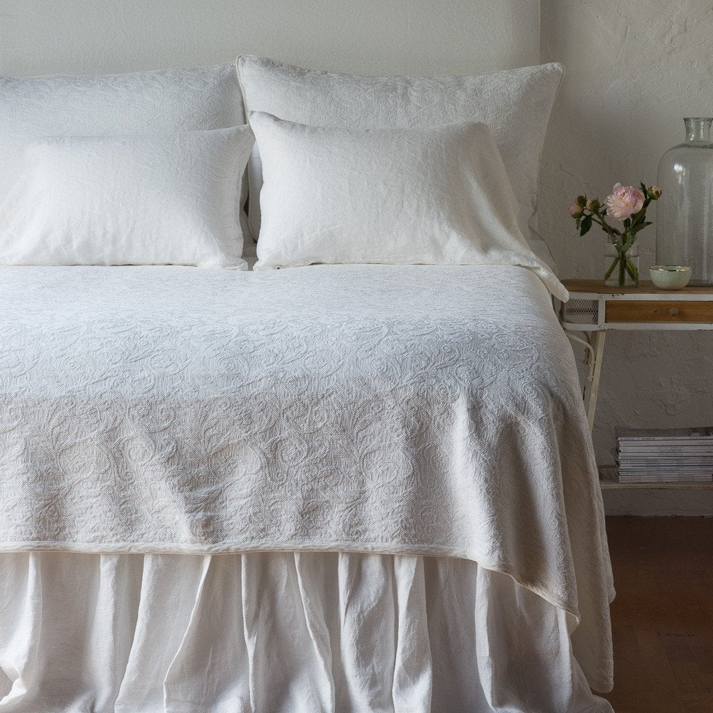 Winter White | The Adele coverlet in winter white, viewed from the foot of the bed, with winter white bed skirt and pillows visible. The headboard is plain light color, and part of a bedside table is visible with flowers, a vase and some books. against a plain white wall.