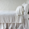 Winter White | The Adele coverlet in winter white, on a bed viewed from the side against a plain white wall. The bed is styled neatly with the coverlet folded back to reveal winter white sheets, bed skirt and pillows.