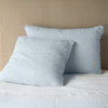 Two Adele shams shown in euro and deluxe sizes, leaning against a neutral toned headboard on white bedding.