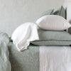 Adele shams in eucalyptus on a bed shown from the side, layered with white sheets and pillowcases and a eucalyptus coverlet against a plain white wall.