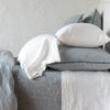 Mineral | Adele shams in mineral on a monochromatic bed shown from the side, layered with white sleeping pillows on sheets and a coverlet, against a plain white wall.
