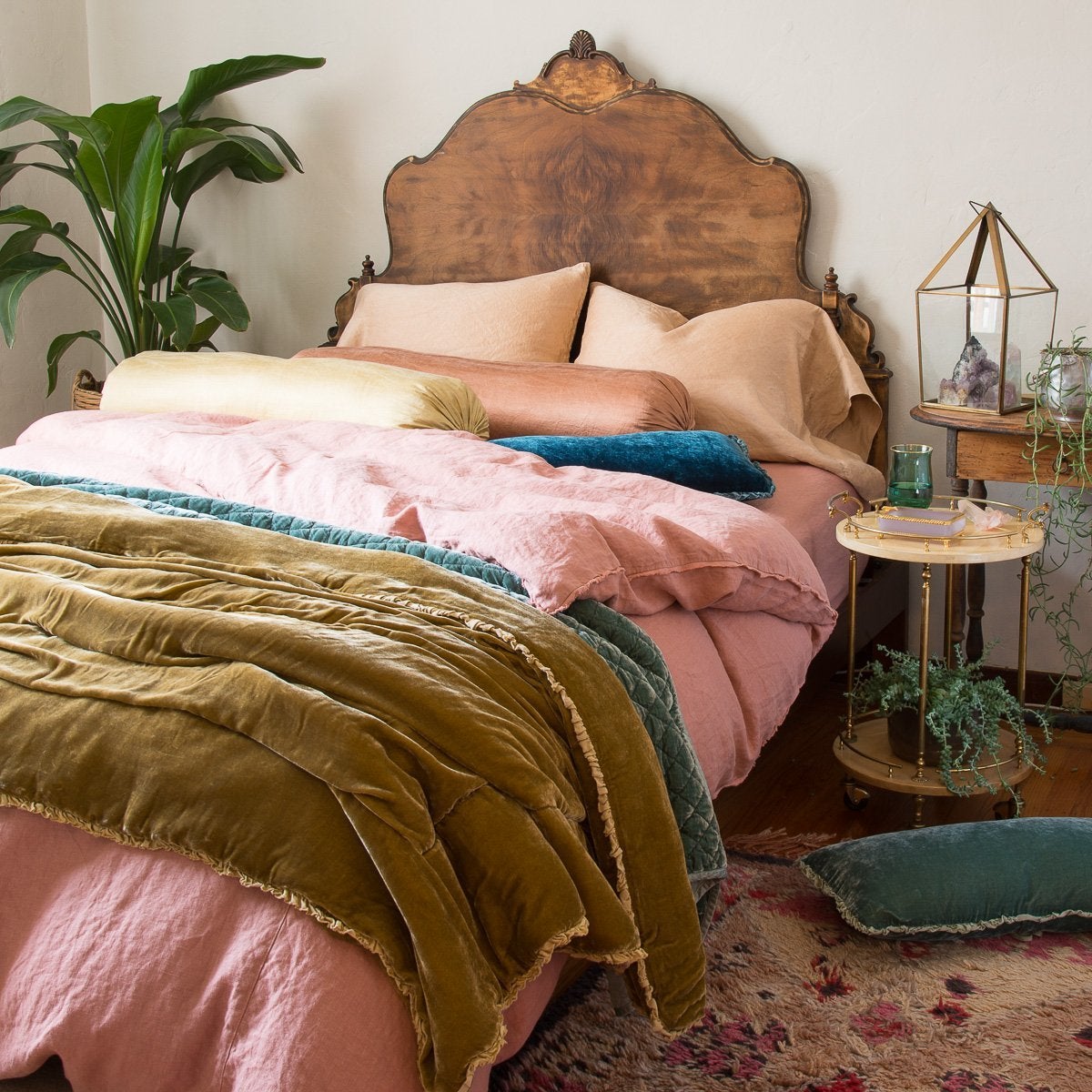 Austin duvet cover shown from three-quarter angle, layered with silk velvet and silk charmeuse accessory pieces in pink, gold and blue-green tones. Dramatic wood headboard, greenery, patterned rug, and eclectic bedside styling create a bohemian vibe.