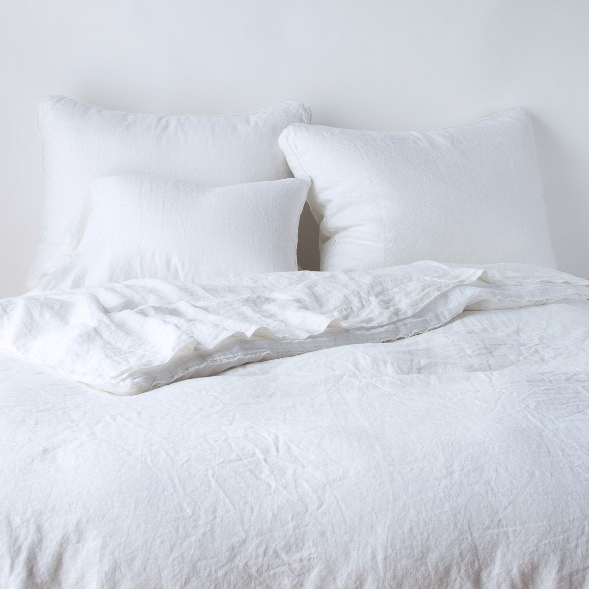 Austin Sham | White | Midweight linen shams shown from foot of bed, leaning against a plain background with monochromatic bedding.
