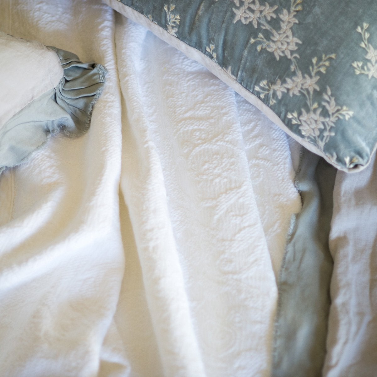 An overhead close up of a rumpled Adele coverlet in white, surrounded by sterling accent pillows and throw blanket.