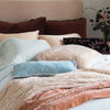 Lynette Blanket | Lynette throw blanket and throw pillow in pearl, layered on rumpled bed with pink, pale blue, and rich brown tones - cropped three-quarter angle.