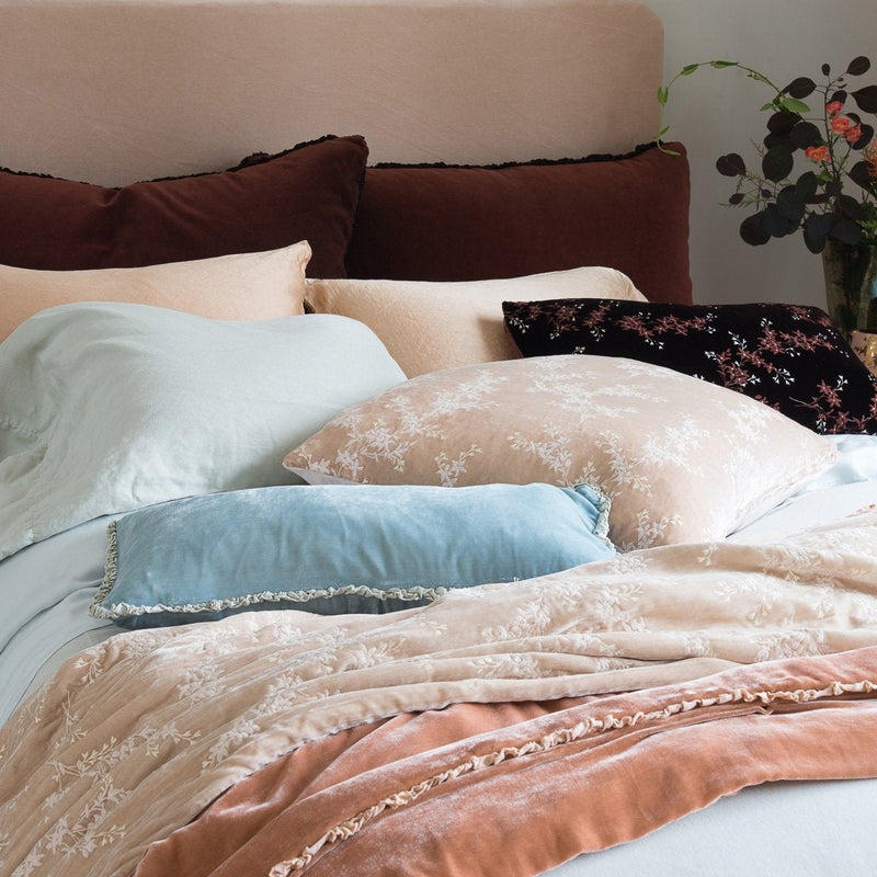 Lynette Blanket | Pearl | Lynette throw blanket and throw pillow in pearl, layered on rumpled bed with pink, pale blue, and rich brown tones - cropped three-quarter angle.